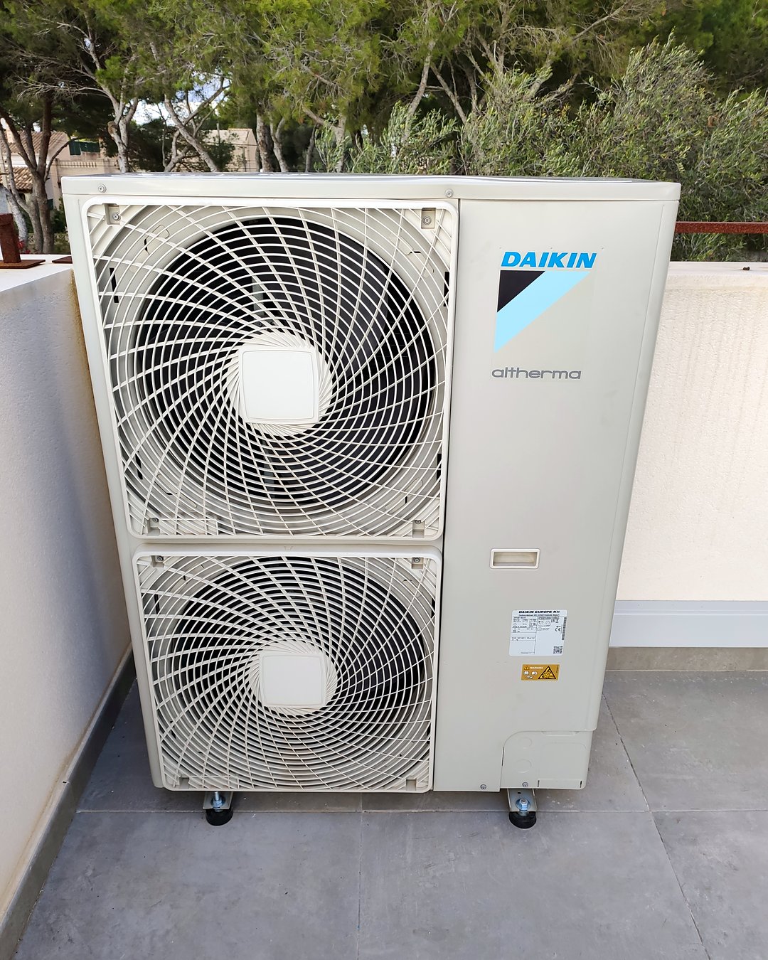 Unidad exterior de bomba de calor Daikin Altherma sobre soportes antivibratorios en una terraza, con pinos detrás, instalada por JSL en Mallorca