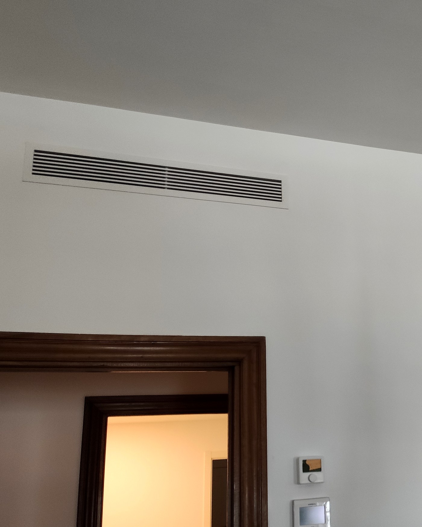 Rejilla lineal de aire por conductos sobre una puerta de madera y termostato en la pared, en una vivienda de Mallorca