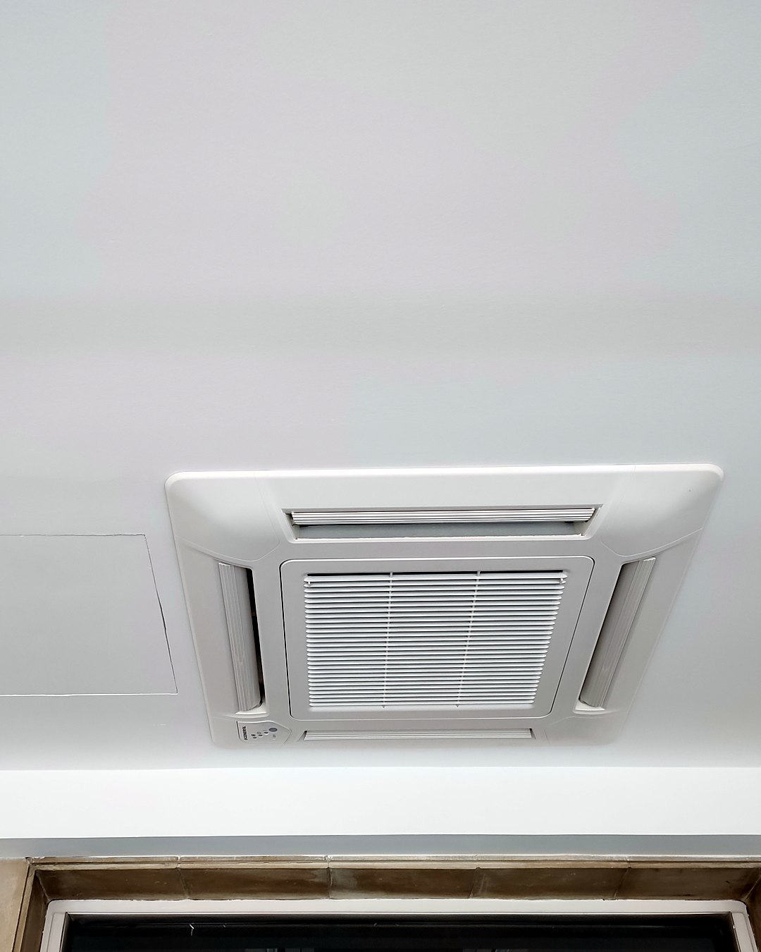 Unidad de aire acondicionado tipo cassette de cuatro vías empotrada en un techo blanco, con su registro al lado, en Mallorca