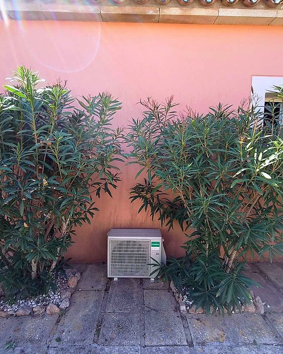 Unidad exterior de aire acondicionado en el suelo de un patio, entre dos adelfas, junto a una pared rosada, una instalación nuestra en Mallorca