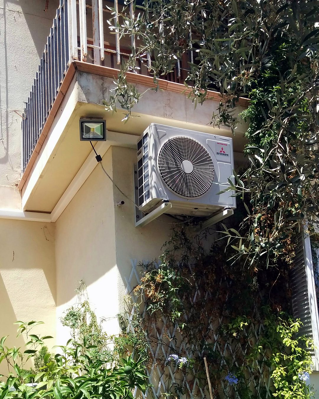 Unidad exterior de aire acondicionado Mitsubishi inverter sobre soporte bajo un balcón, entre plantas trepadoras, en una casa de Mallorca