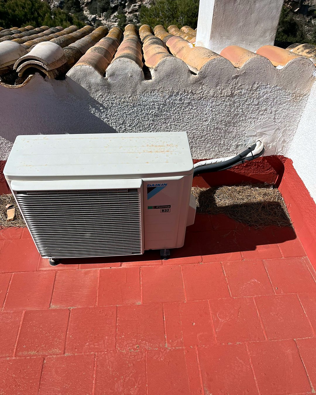 Unidad exterior de aire acondicionado Daikin inverter en una terraza junto a un murete encalado con remate de teja árabe y la ladera detrás, en Mallorca