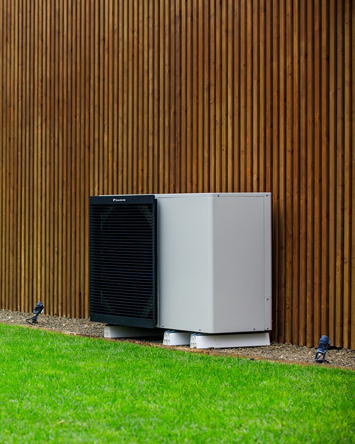Unidad exterior de una bomba de calor Daikin Altherma 3 M junto a una fachada de madera y un jardín. Imagen del fabricante Daikin; no es una obra de JSL