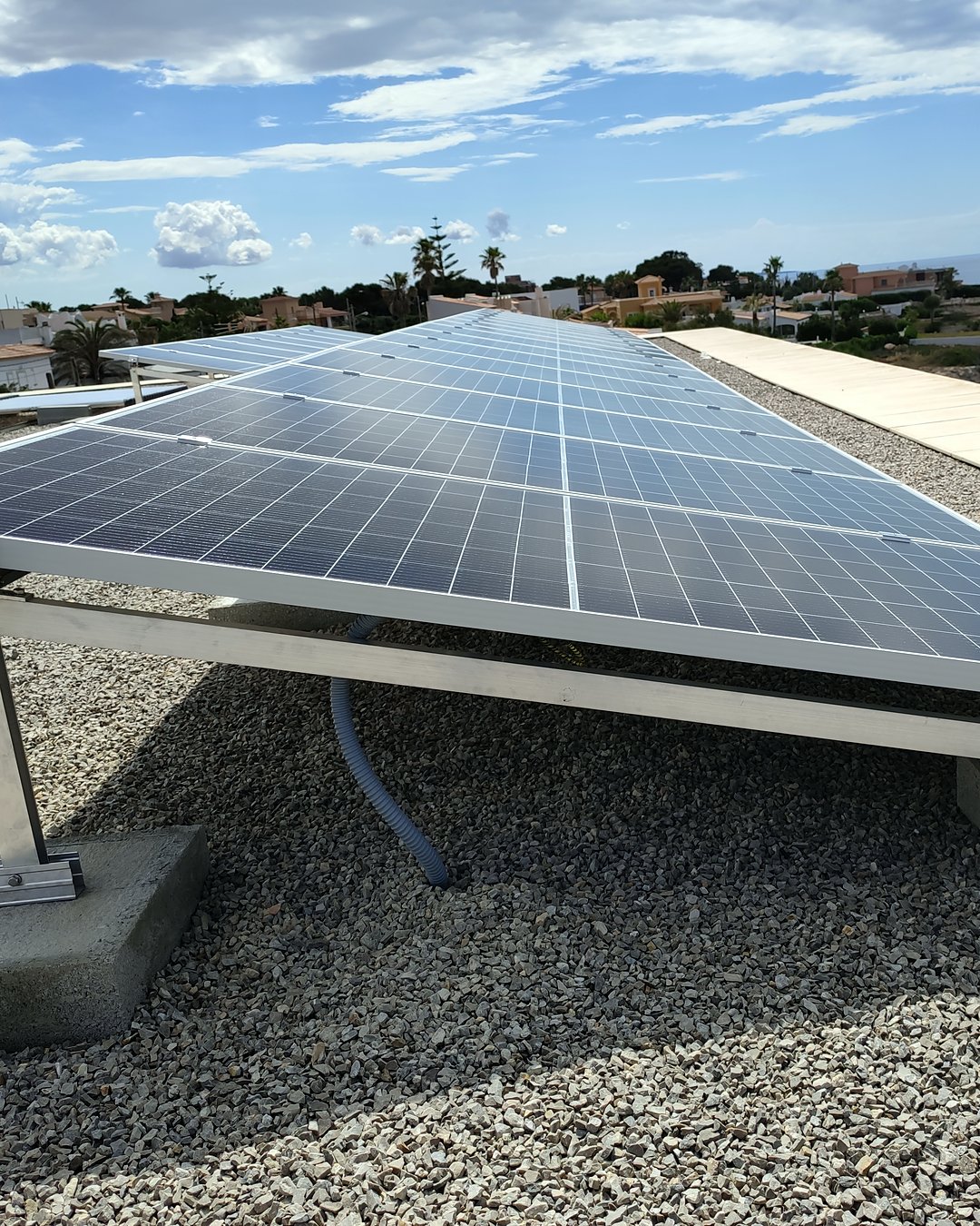 Fila de placas solares inclinadas con lastres de hormigón en una azotea de grava, con chalets entre pinos al fondo, montadas por JSL en Mallorca