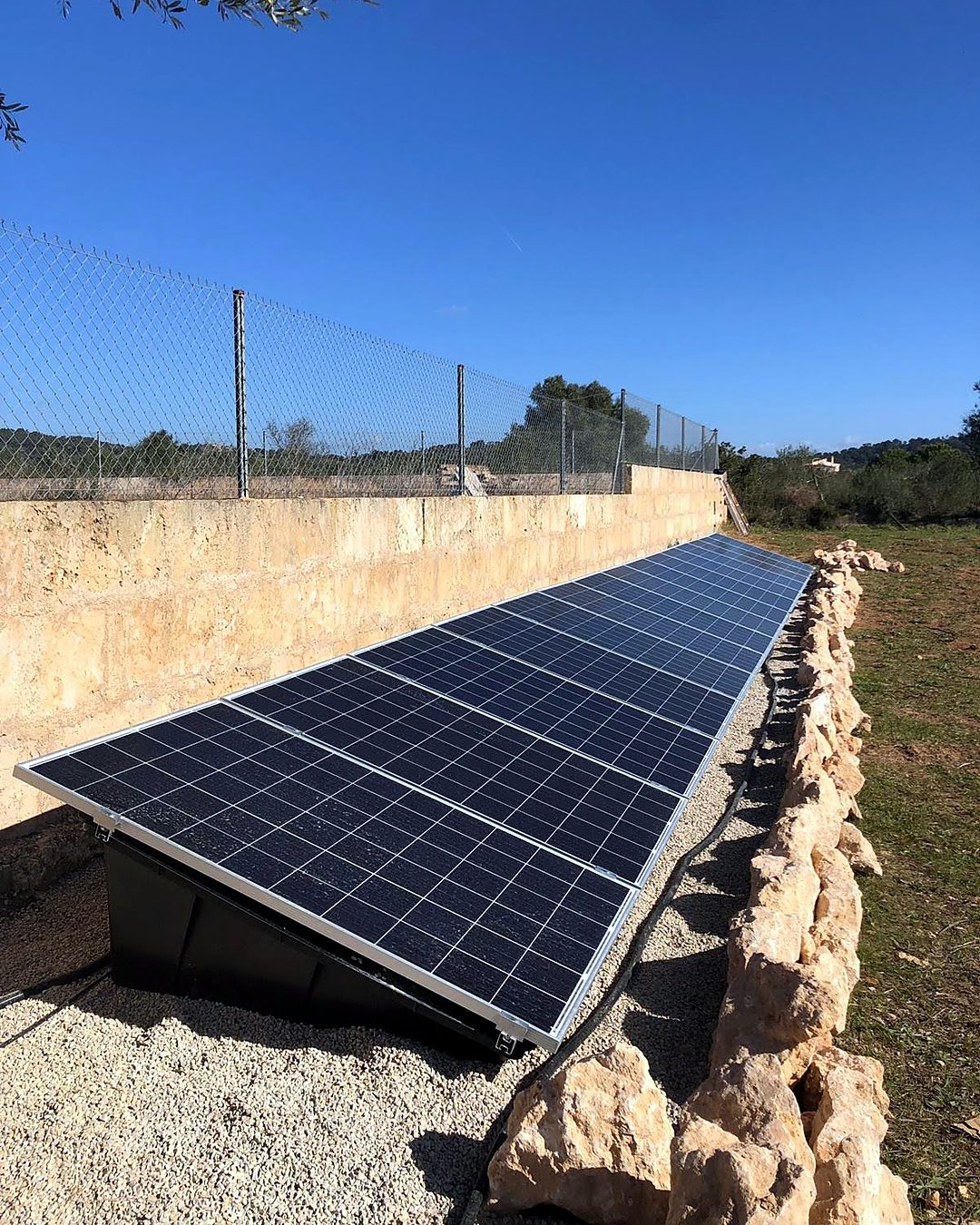 Hilera de placas solares sobre soportes en el suelo junto a un muro de marès y un margen de piedra en una finca de Mallorca, montada por JSL