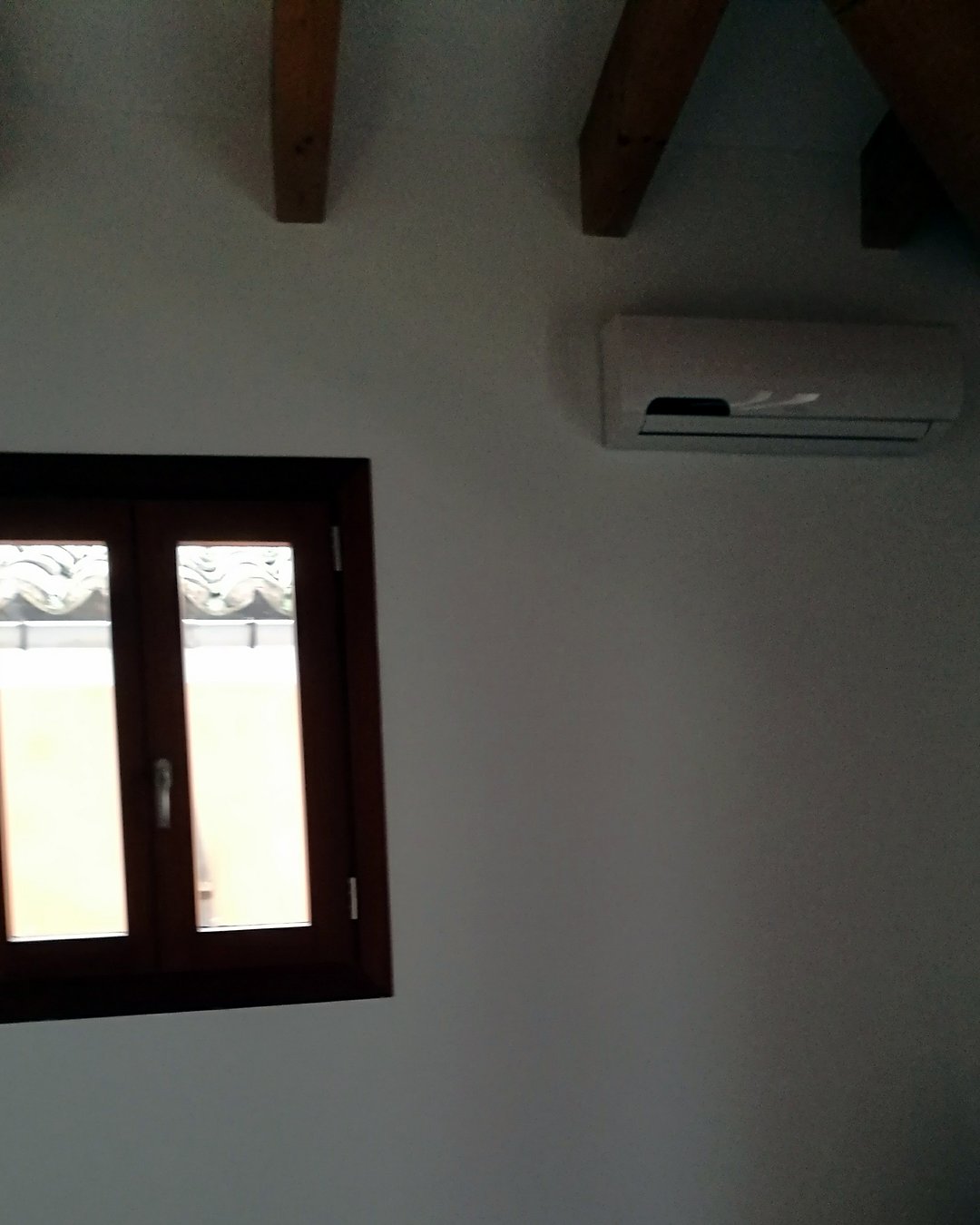 Split de aire acondicionado en lo alto de una habitación con vigas de madera vistas y ventana de madera, montado por JSL en Mallorca